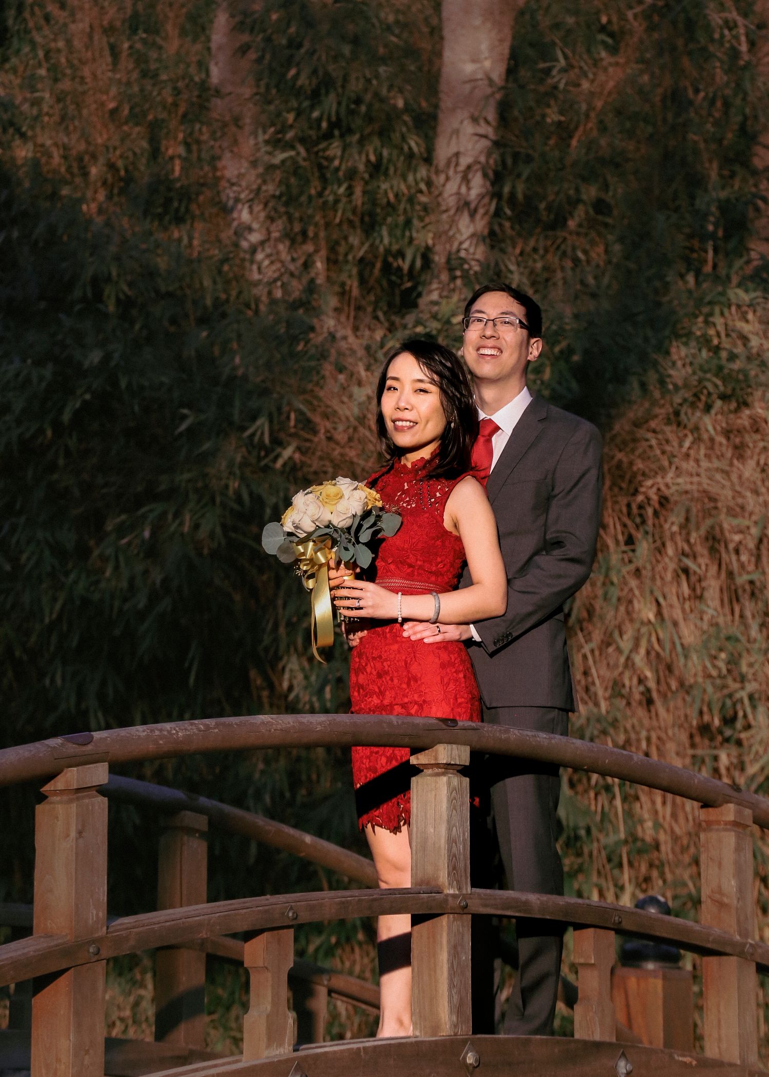 Couple in formal wear poses on wooden bridge during outdoor photoshoot.