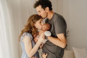 A tender family moment as parents hold and embrace their newborn baby against a soft white curtain background.