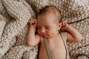 Peaceful sleeping infant resting on soft knitted beige blanket while wearing gray suspenders.