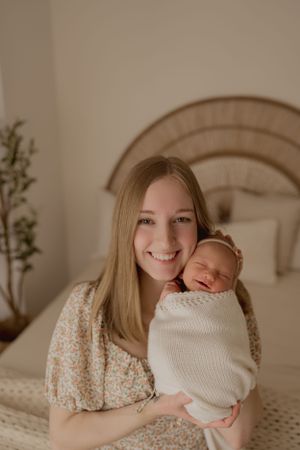 Mother in floral dress holds sleeping newborn wrapped in white blanket while sitting on bed with rattan headboard.