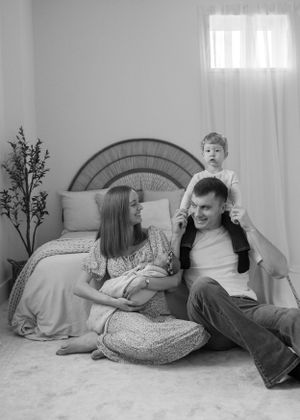 A candid black and white family photo showing tender moments between family members lounging by a bed with rattan headboard.
