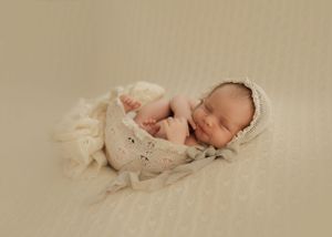 Peaceful sleeping newborn swaddled in a cream colored wrap lying on a soft neutral backdrop.