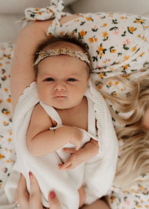 A small baby wearing a lace headband lies on a white blanket against floral patterned fabric.