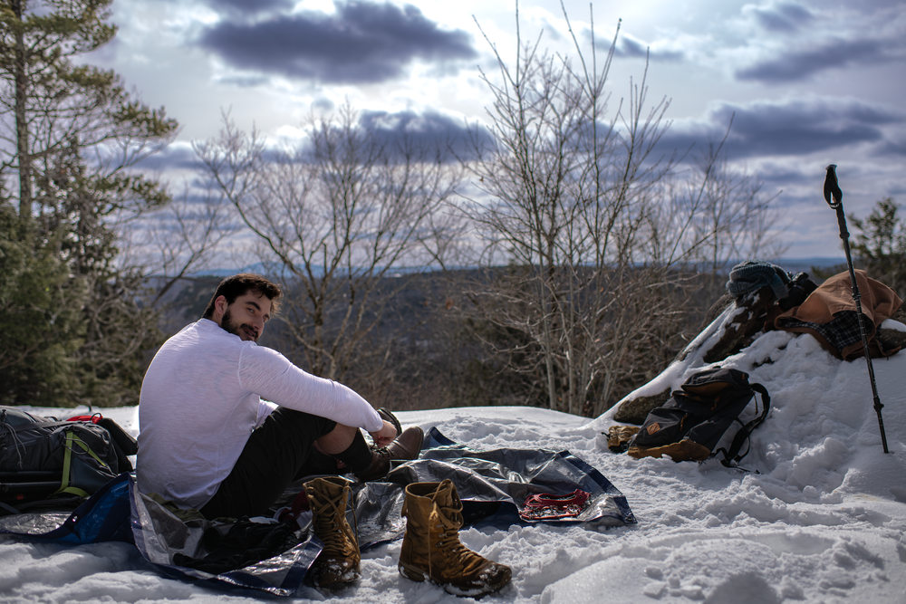 A person in winter clothing sits near a campfire in the snow while camping with mountain views in the background.