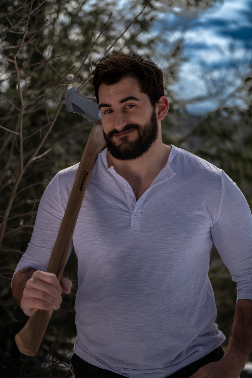 Bearded person wearing a white henley shirt holding an axe while standing outdoors with trees and blue sky in background.