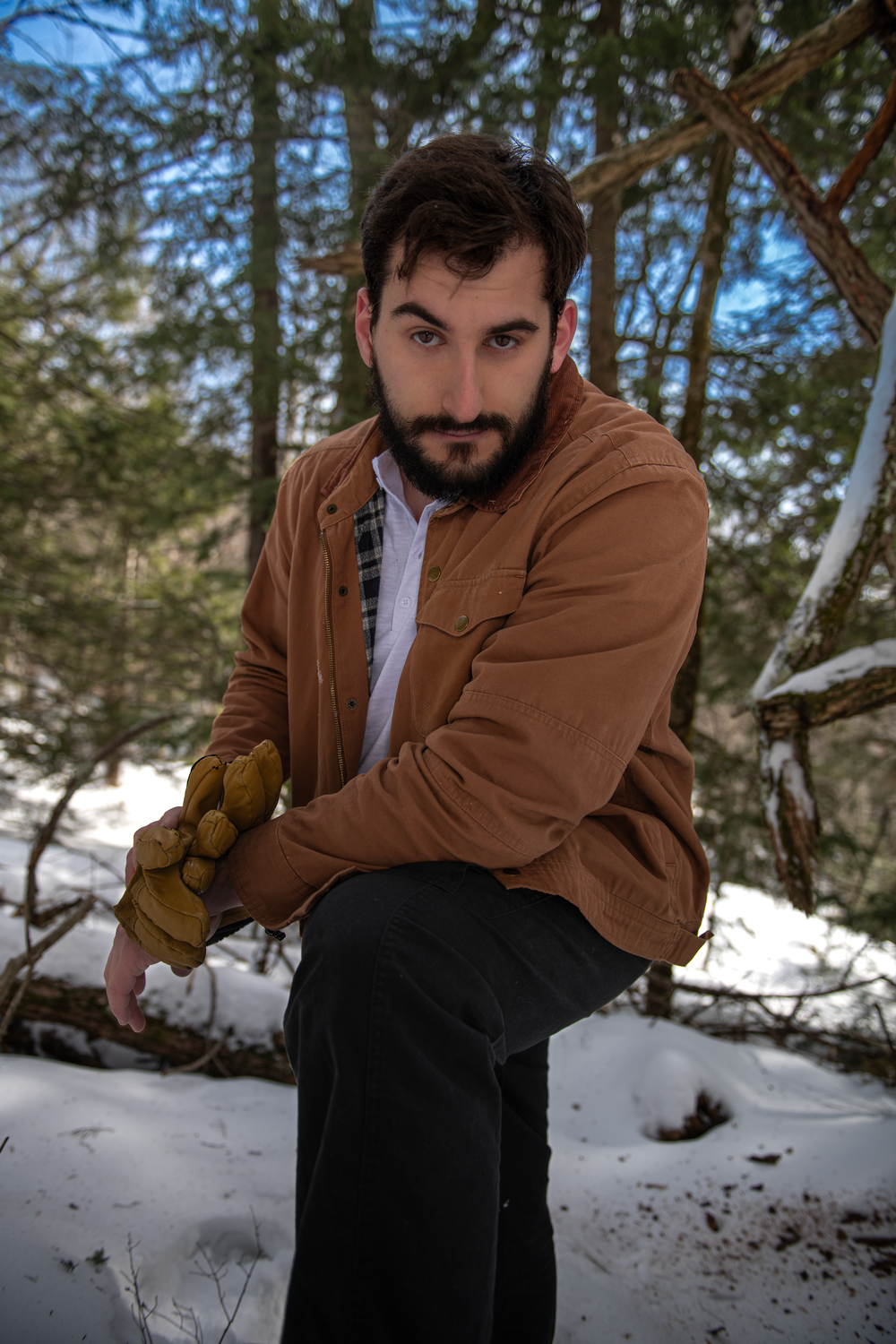 A person in a brown jacket and work gloves poses in a snowy forest setting.