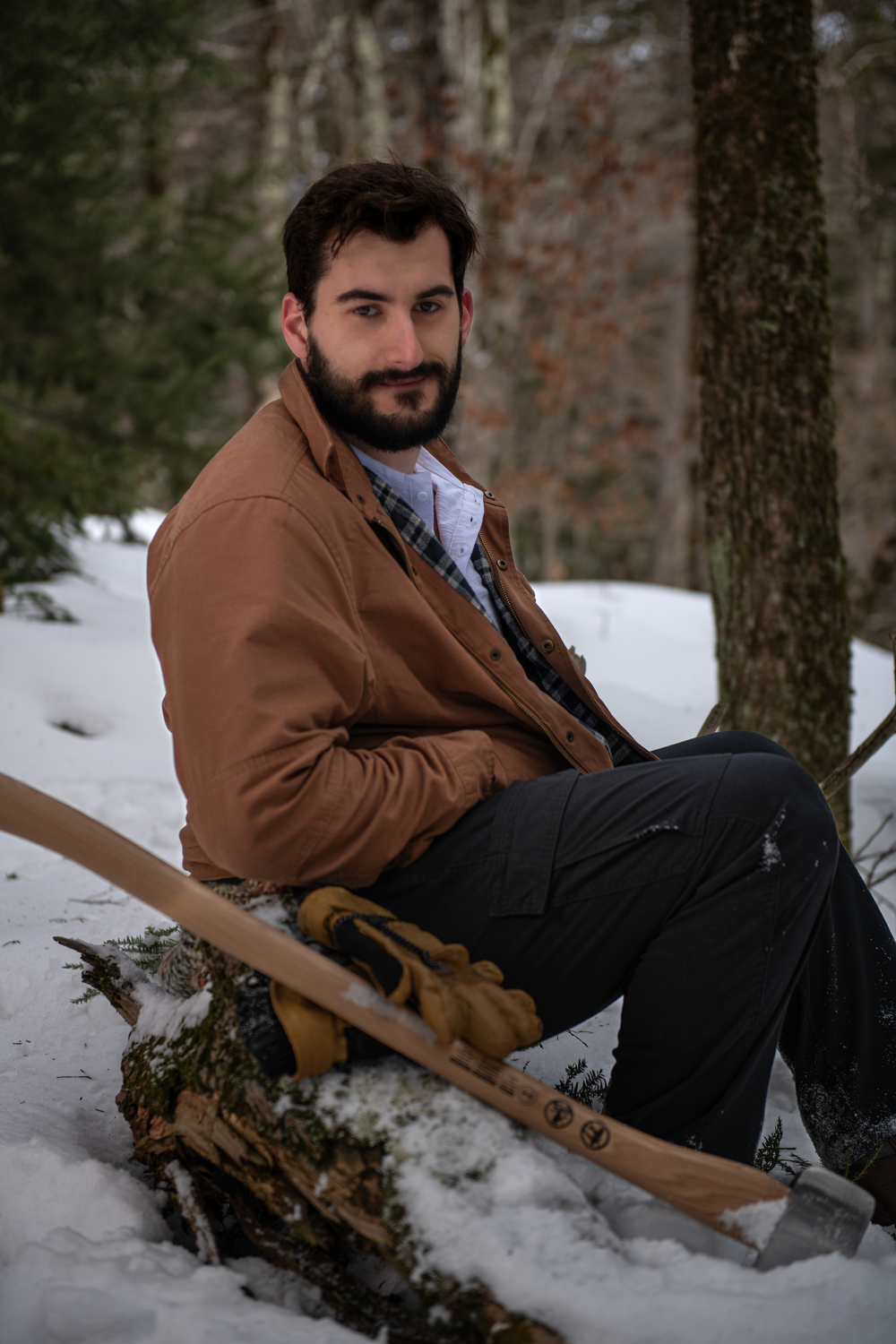 Person in brown jacket sits on snowy log in winter forest with axe and work gloves.