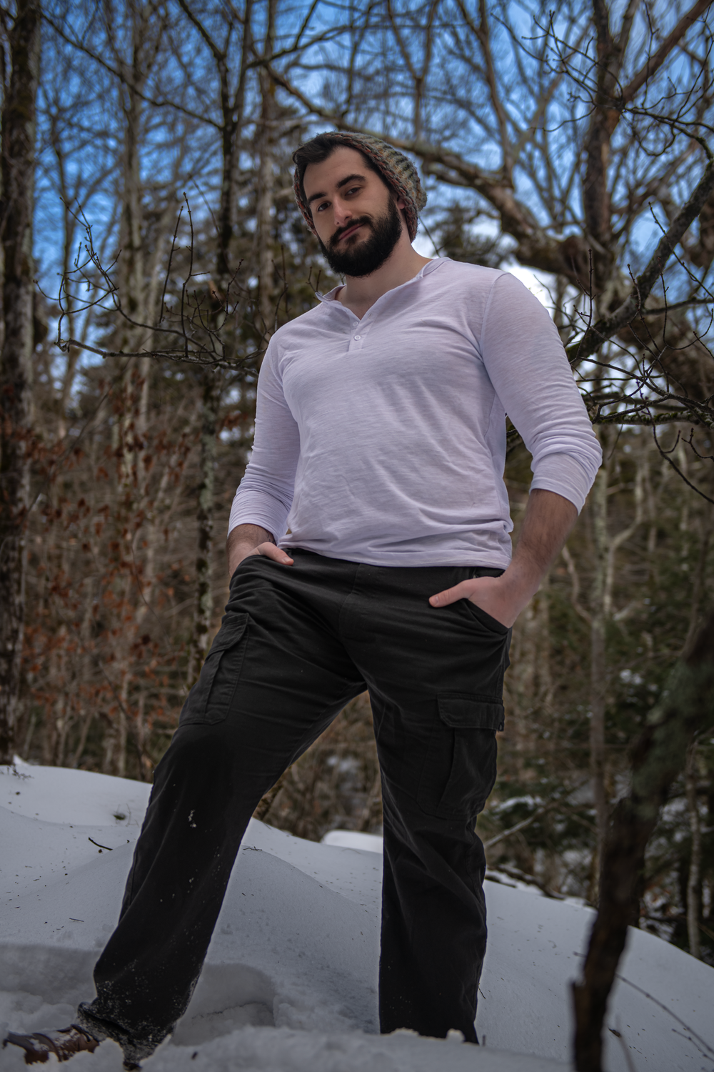Person in a white henley shirt and dark cargo pants poses in a snowy wooded setting during winter.
