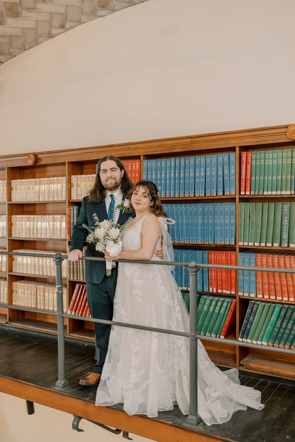 A wedding couple poses together on a library mezzanine with floor-to-ceiling bookshelves in the background.