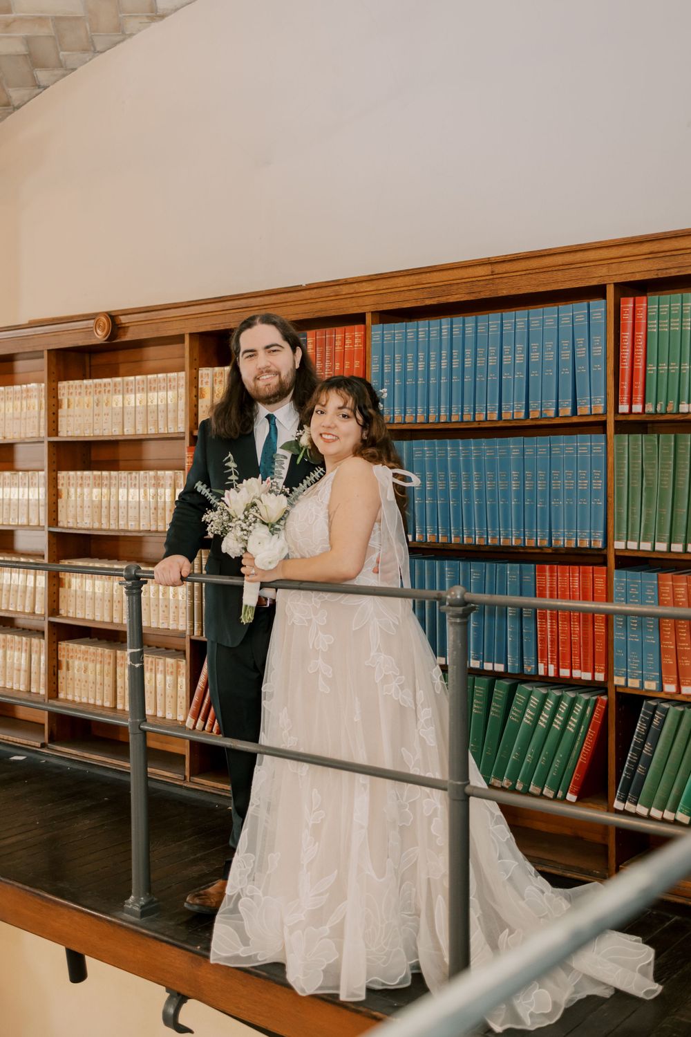 A wedding couple stands against a backdrop of colorful library books in a traditional library setting.