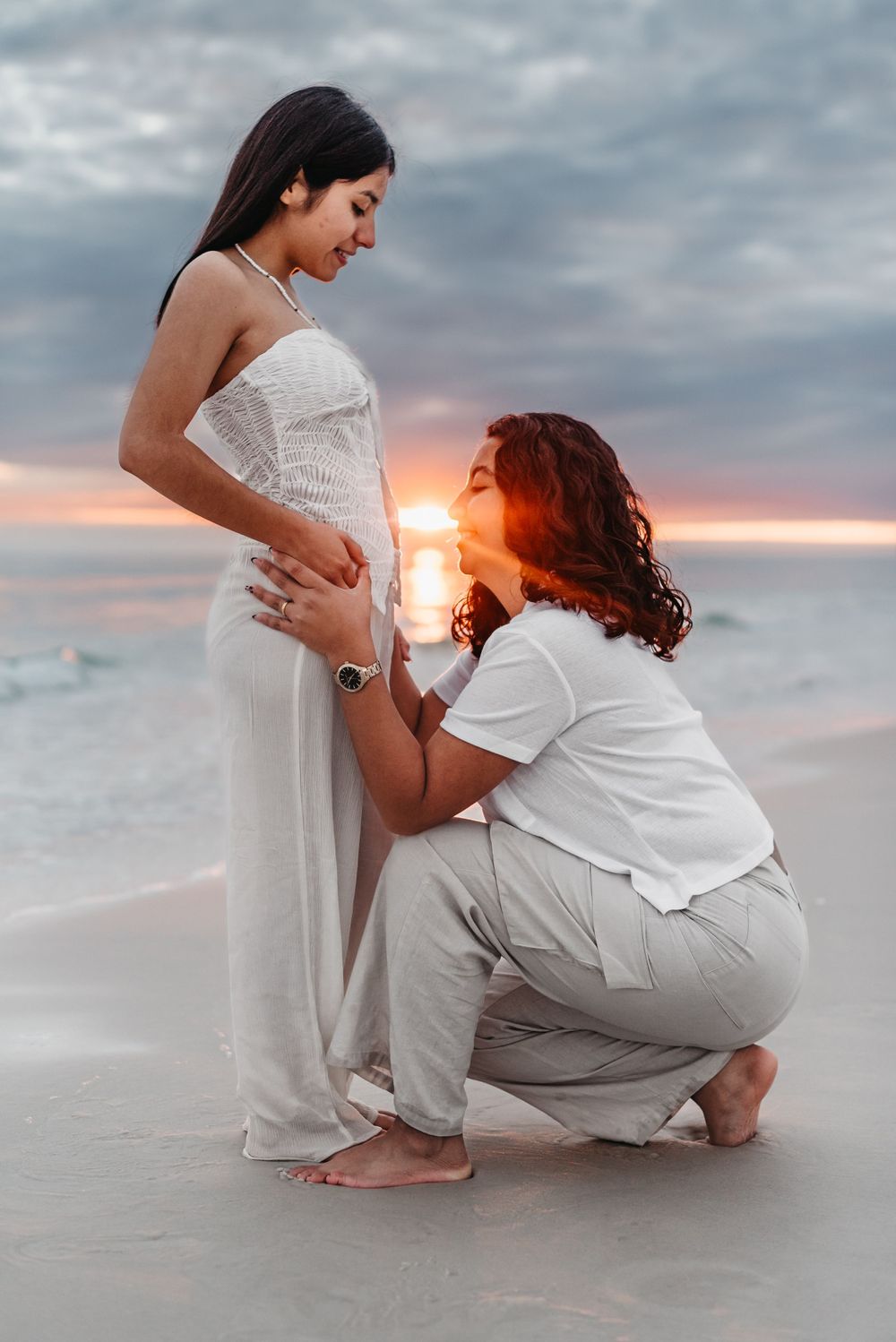 Two people in white outfits sitting close together on a beach at sunset.
