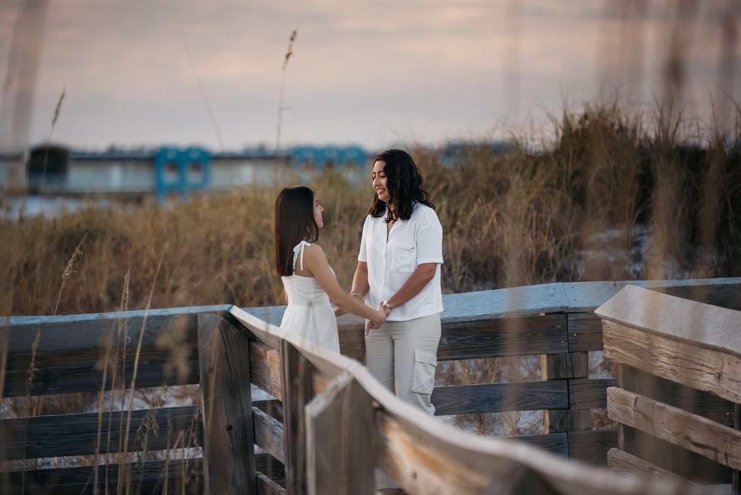 A couple stands together on a wooden boardwalk surrounded by tall beach grass at dusk.
