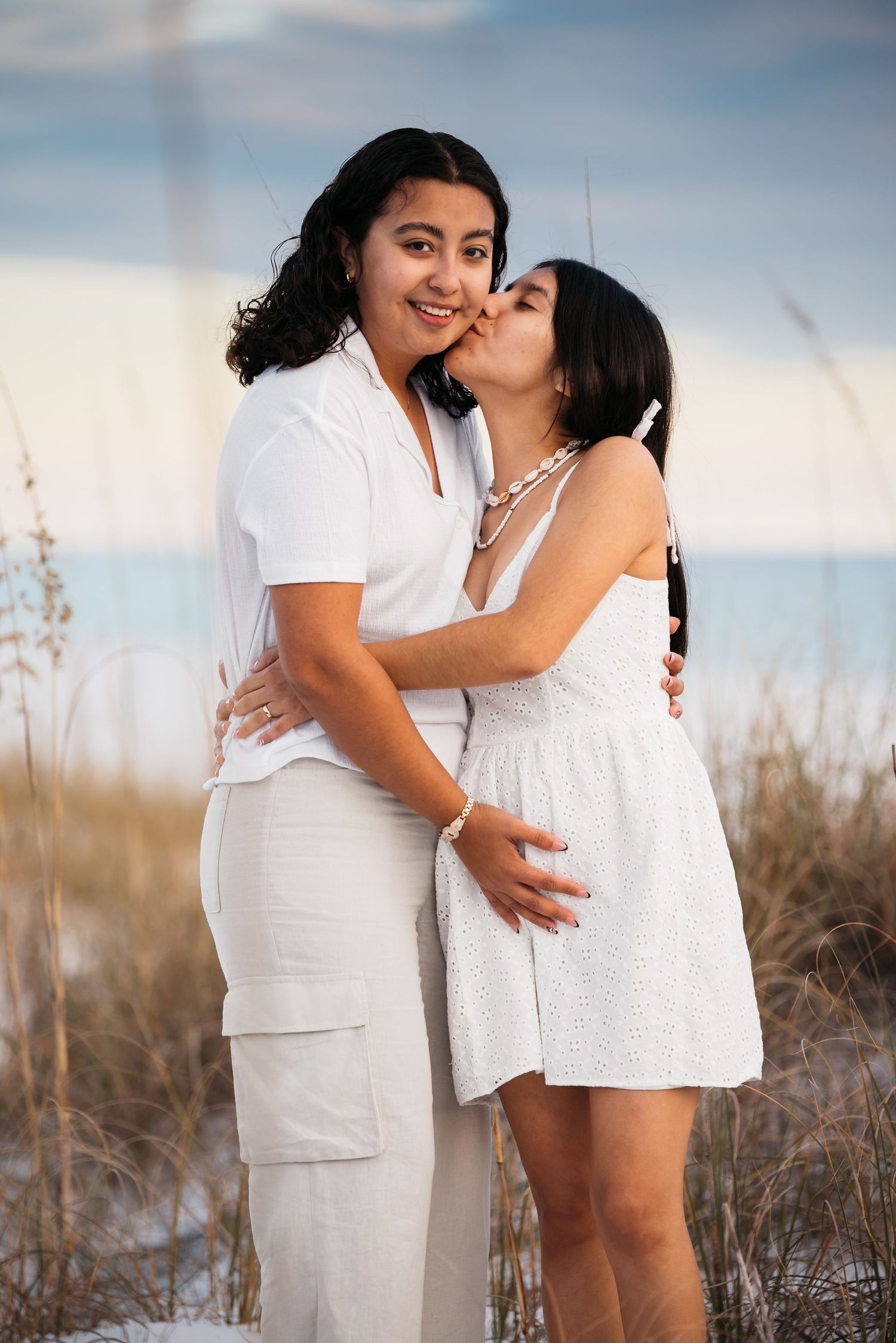 A couple in white outfits share an intimate moment on the beach at sunset.