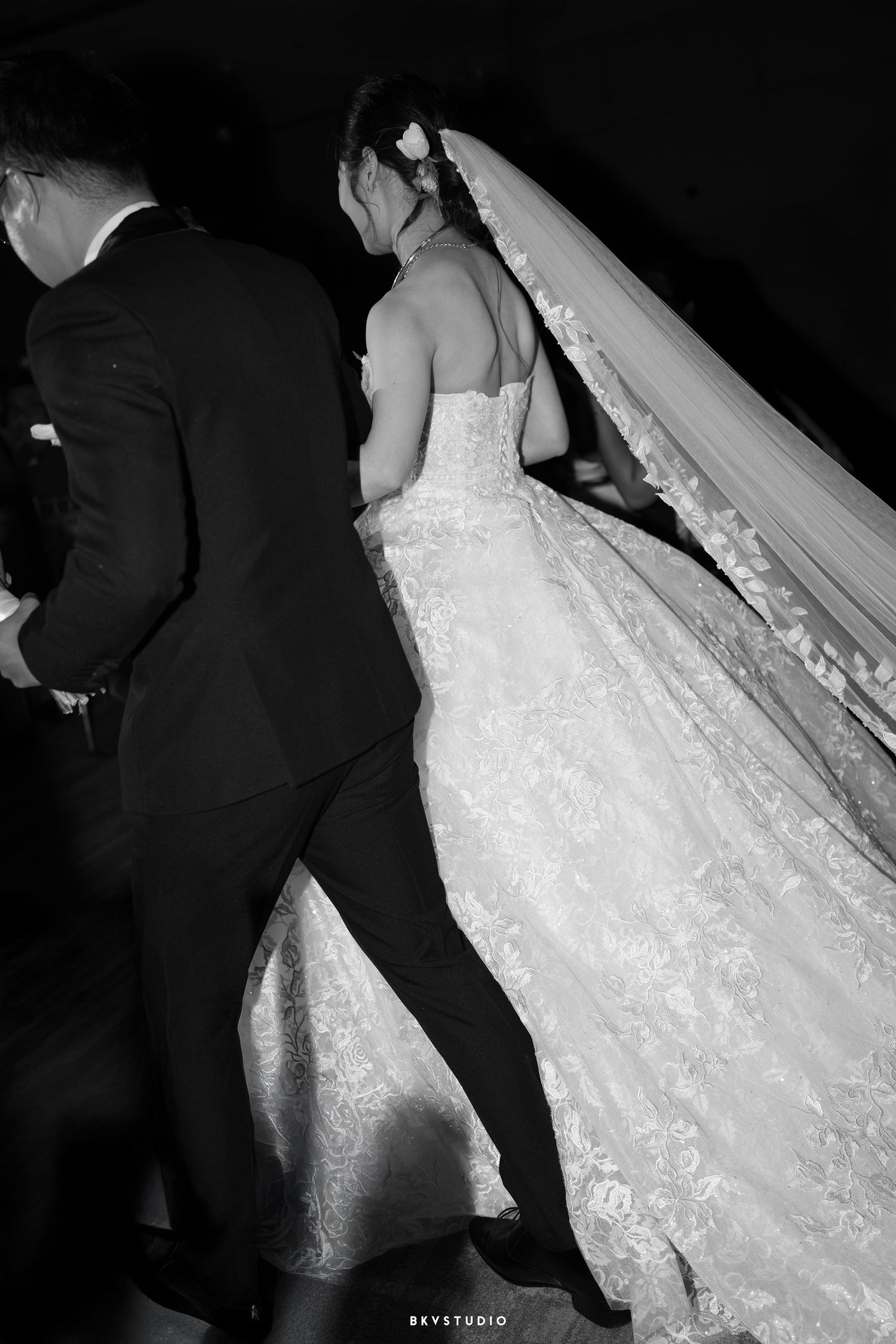 Black and white wedding photo showing the back of a bride's detailed lace gown with flowing veil next to a suited figure.