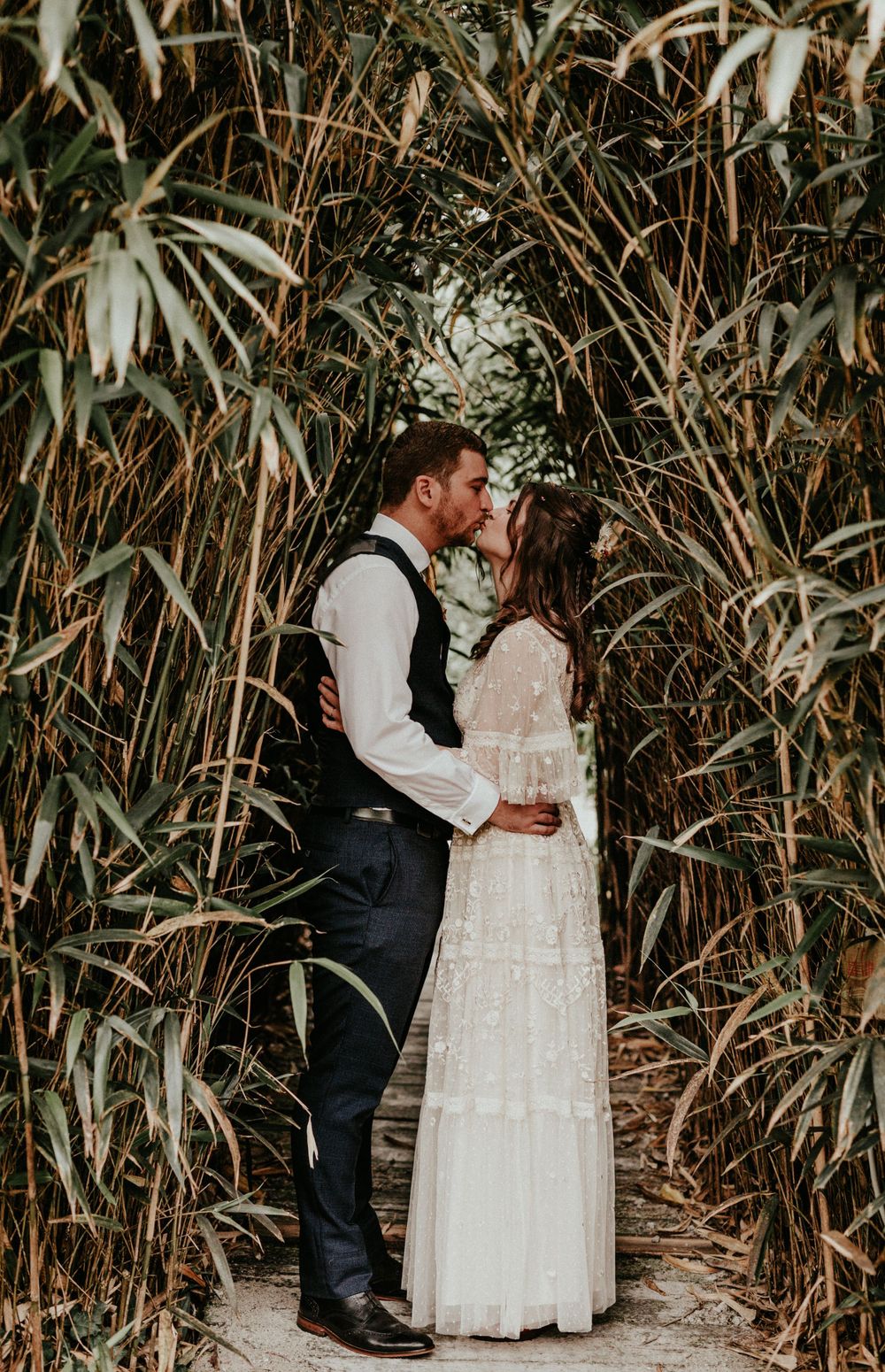 A couple shares a romantic kiss surrounded by tall bamboo plants on a narrow pathway.