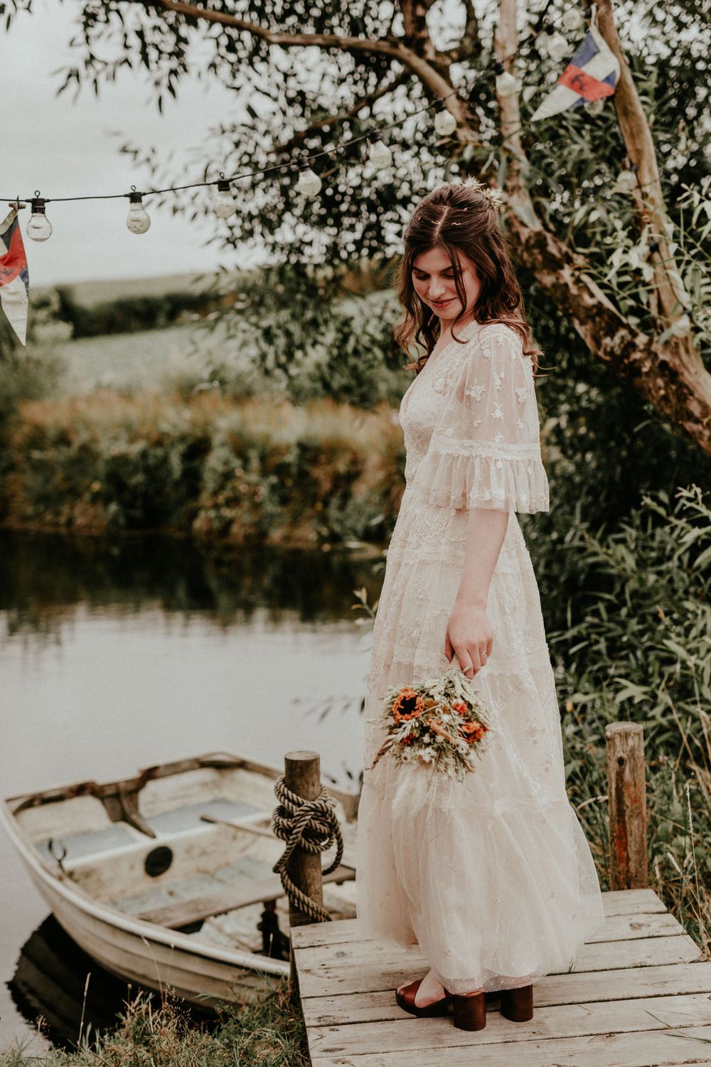 A person in a flowing white vintage-style dress stands on a wooden dock by a small boat at sunset holding dried flowers.
