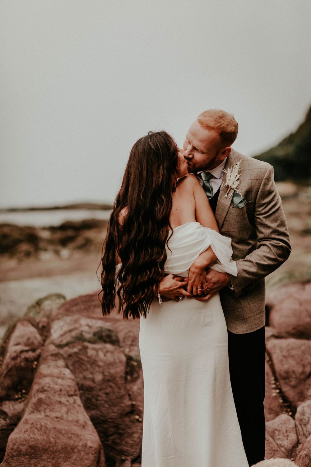 A romantic wedding couple share an intimate moment on rocky coastal cliffs at dusk.