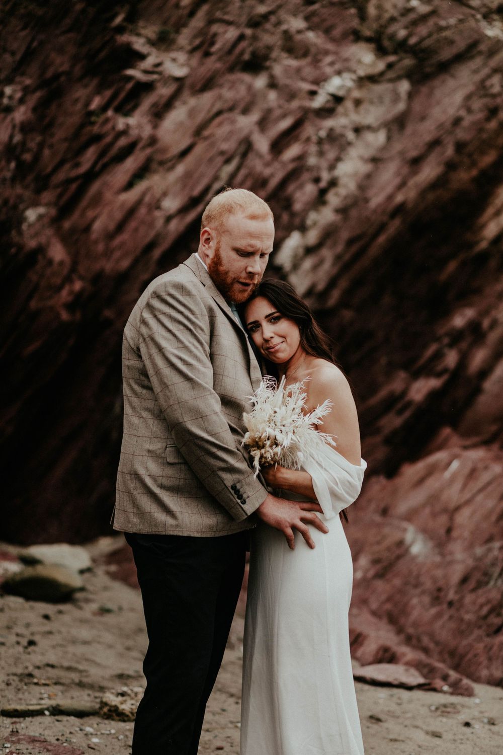 A couple in elegant wedding attire embrace tenderly against a dramatic red rock cliff background.