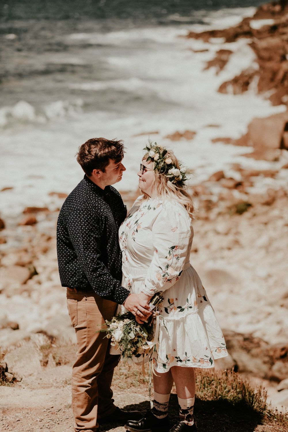 A couple in boho wedding attire stands together on a rocky cliff overlooking a misty landscape.