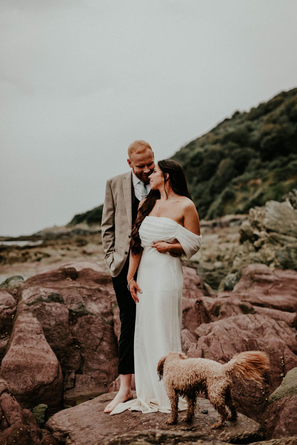 Couple shares an intimate moment on rocky coastal landscape with their fluffy dog beside them at sunset.