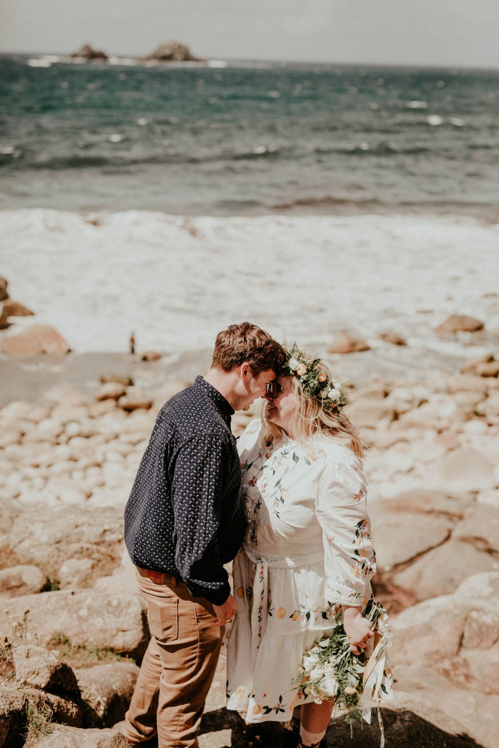 A romantic coastal scene with two people embracing on rocky shores with crashing waves and ocean views in the background.