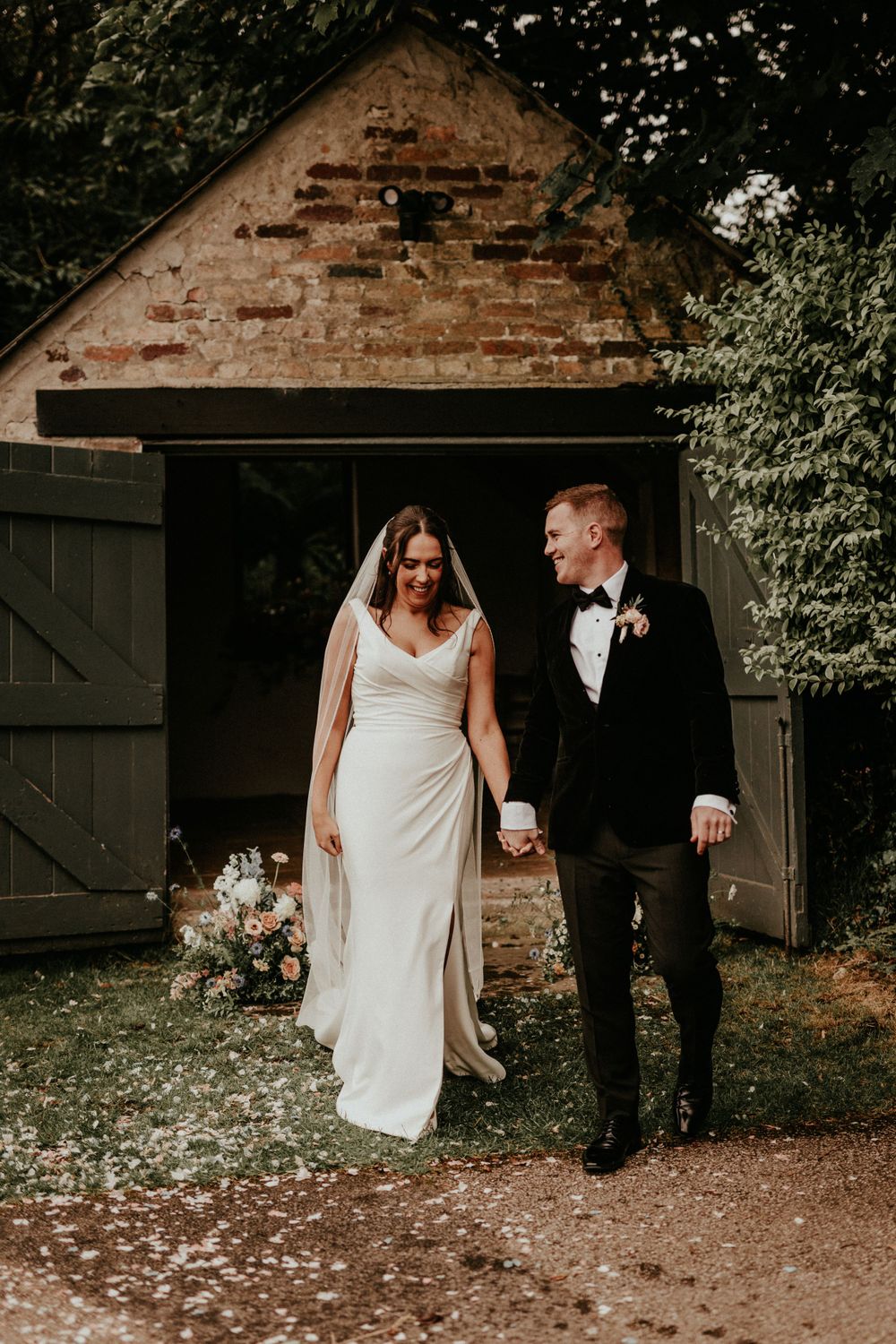 Newlywed couple emerges from rustic barn doorway in romantic wedding photo with exposed brick and greenery backdrop.