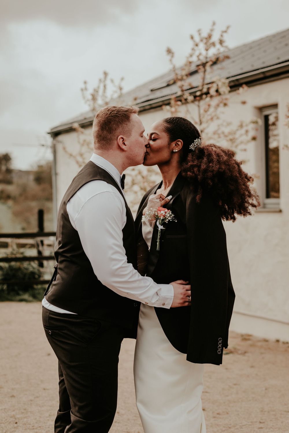 A romantic wedding couple sharing a kiss outdoors during their vintage-style photoshoot near a rustic building.