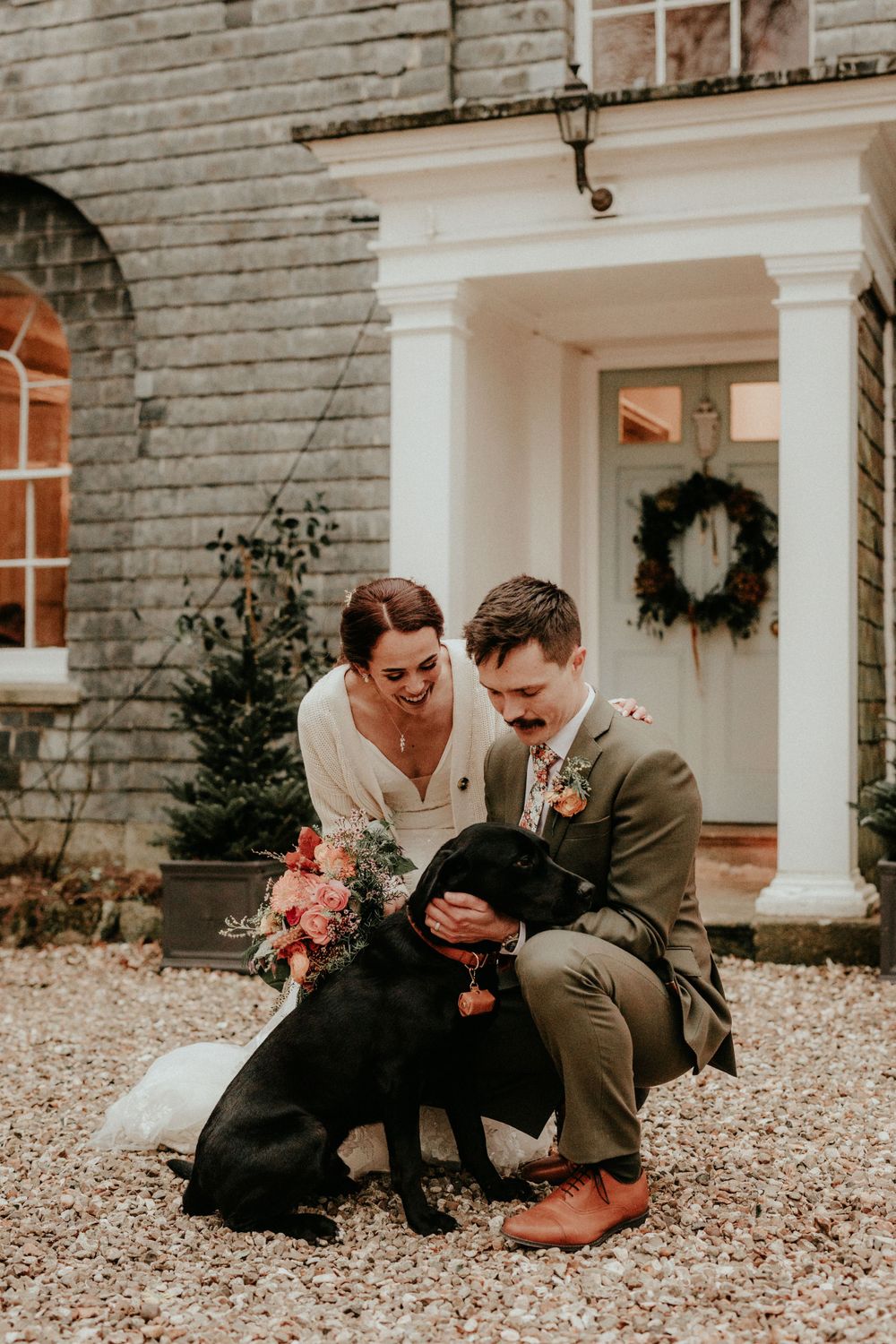 Couple in wedding attire kneeling on gravel driveway with their black Labrador dog outside brick home with wreath.