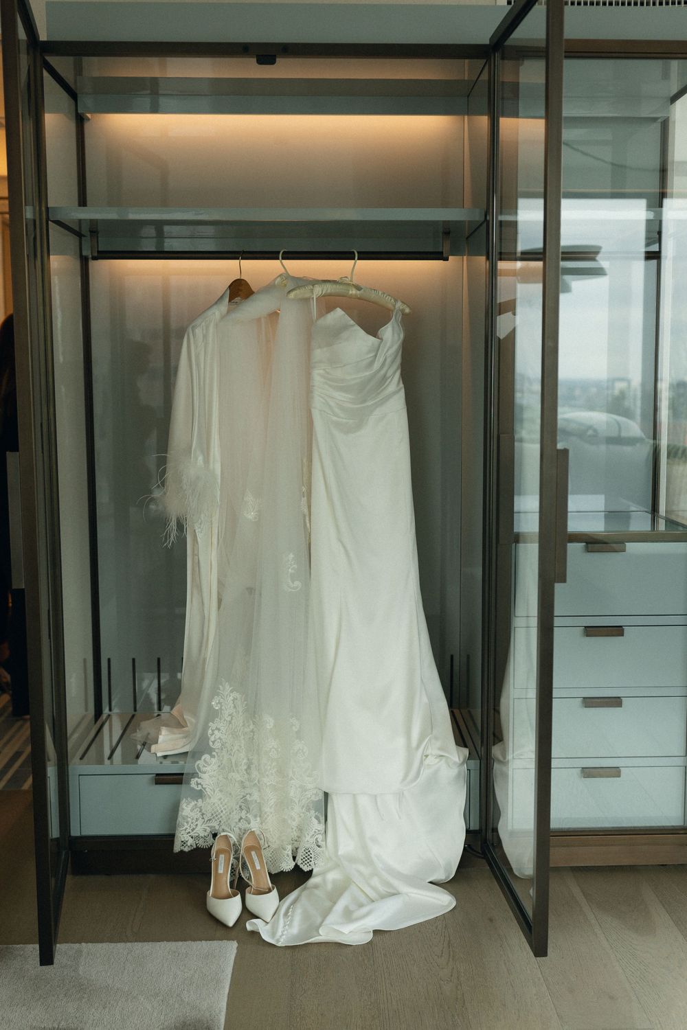Elegant white wedding dress and veil hanging in a modern glass display wardrobe with matching shoes beneath.