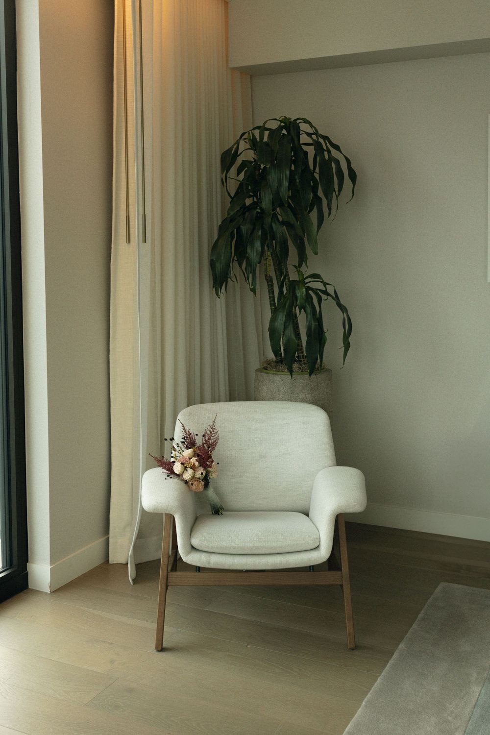 Modern white armchair with wooden legs sits beside tall indoor plant and window in minimalist interior setting.