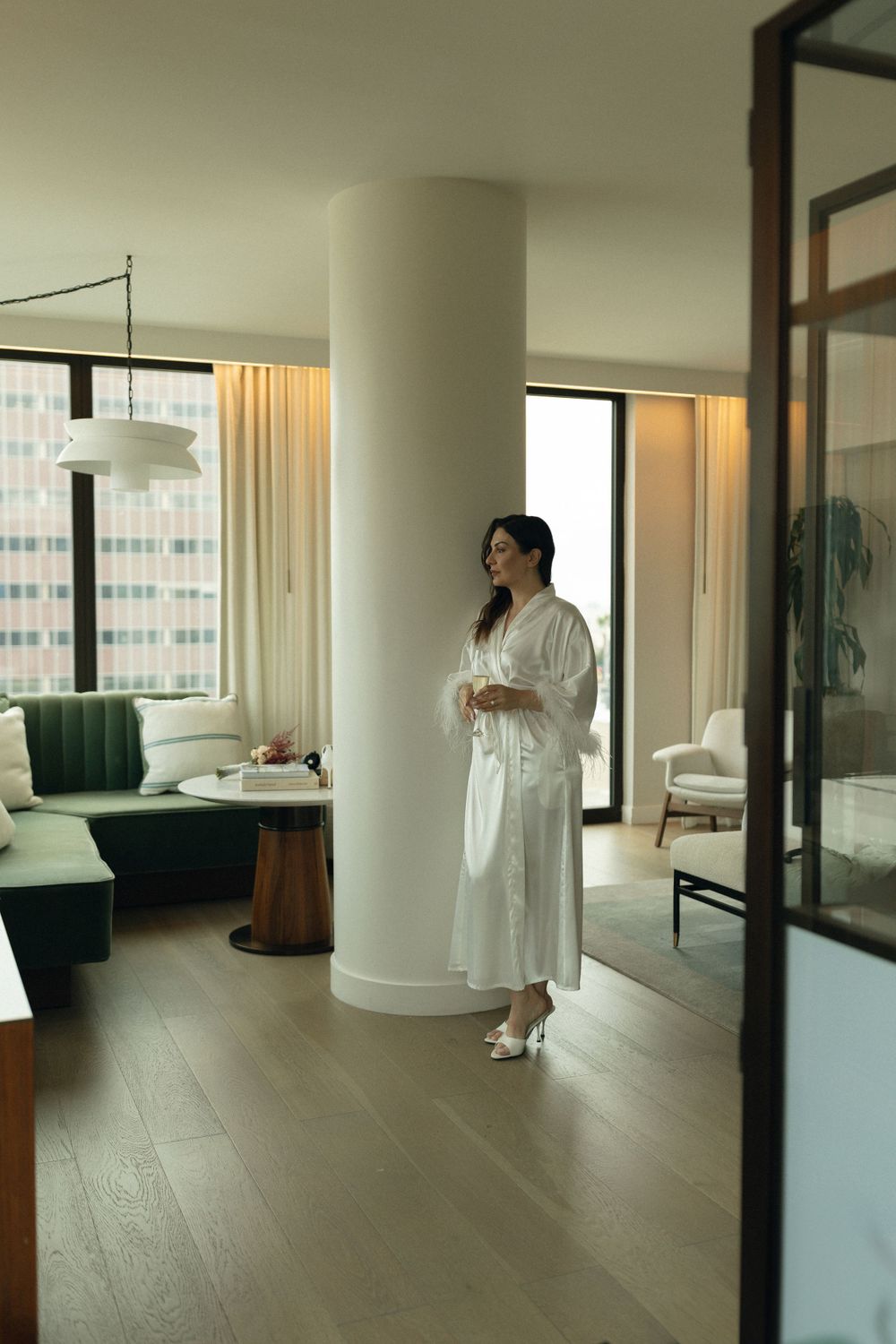 Someone in a white bathrobe stands near large windows in a modern apartment with hardwood floors and elegant furniture.