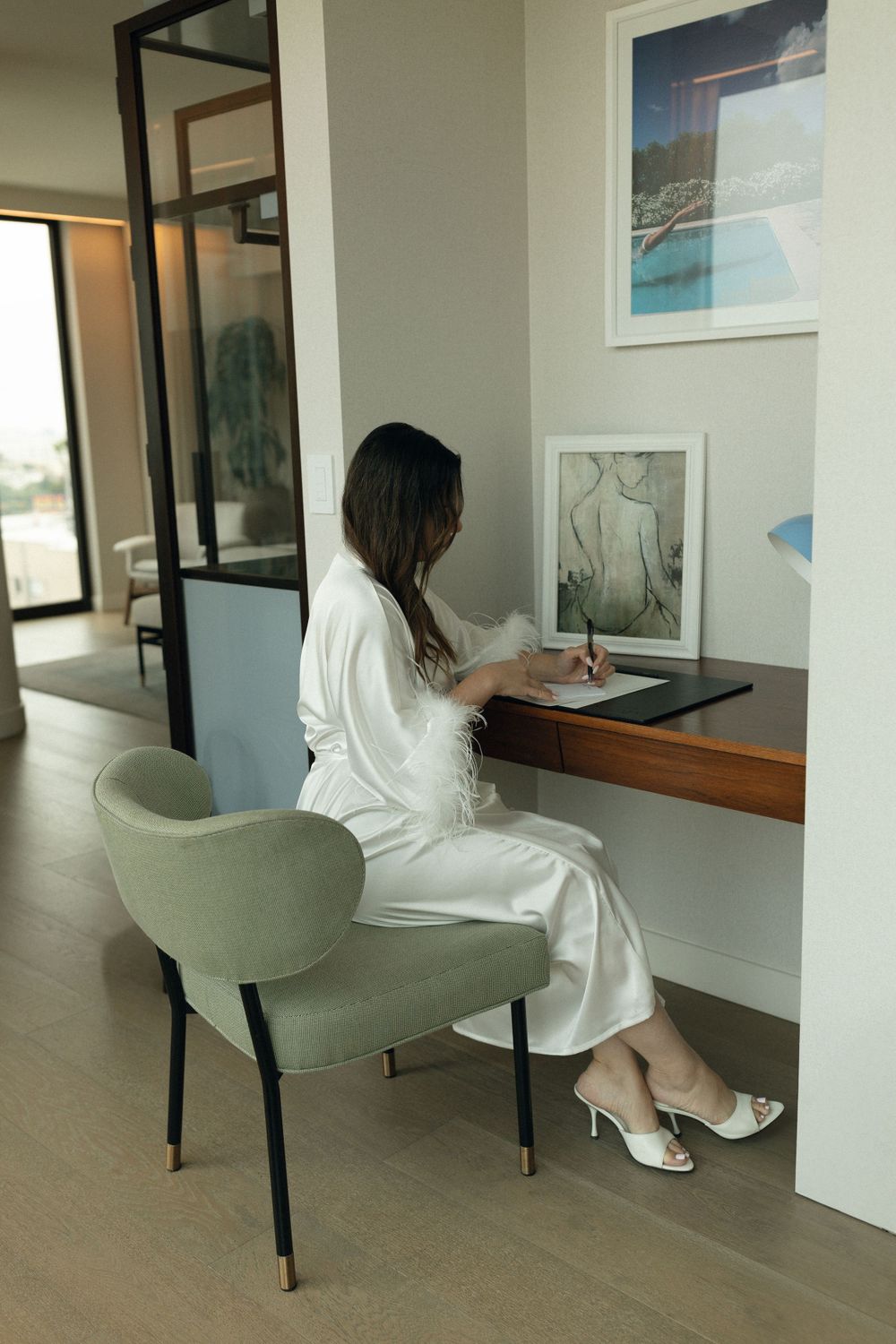 Person in white outfit sits at wall-mounted desk with modern mint green chair working on laptop in minimalist space.