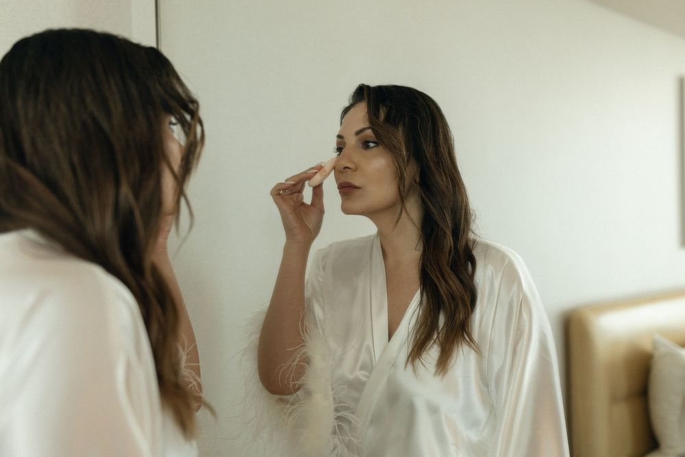A person with long brown hair applies lip gloss in front of a bright window.