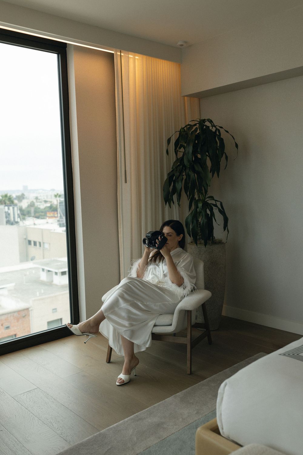 Person in white robe lounges by window in minimalist room with indoor plants and natural light.