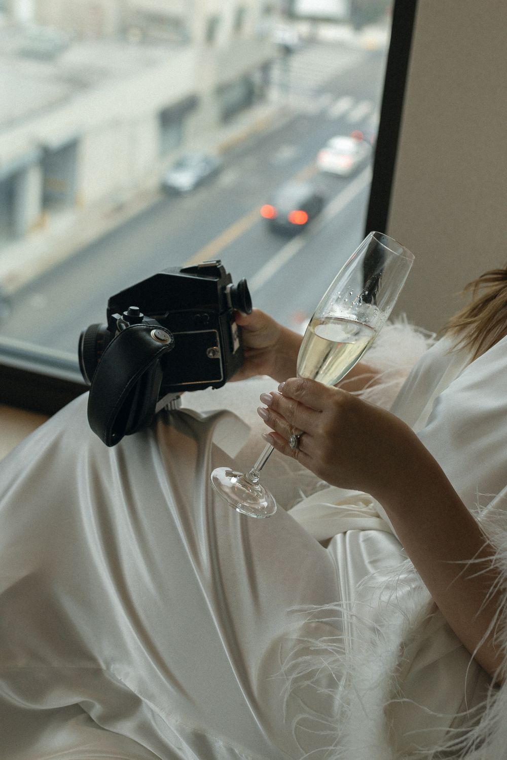 Person in white robe holding DSLR camera and champagne glass near window overlooking city street.