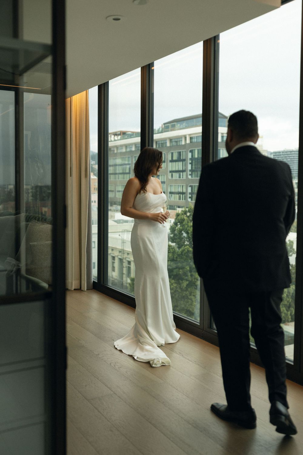 A couple stands by floor-to-ceiling windows overlooking a city view in formal wedding attire.