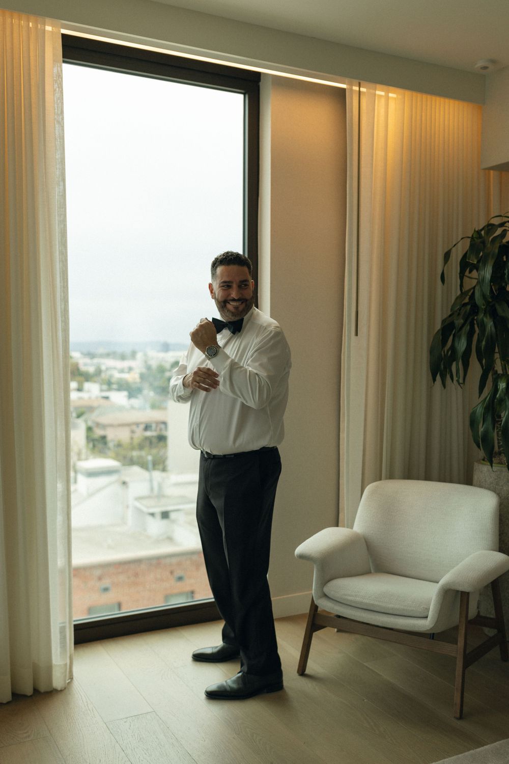 A person adjusts their cuffs while standing near a large window in an elegant room with white curtains and modern decor.