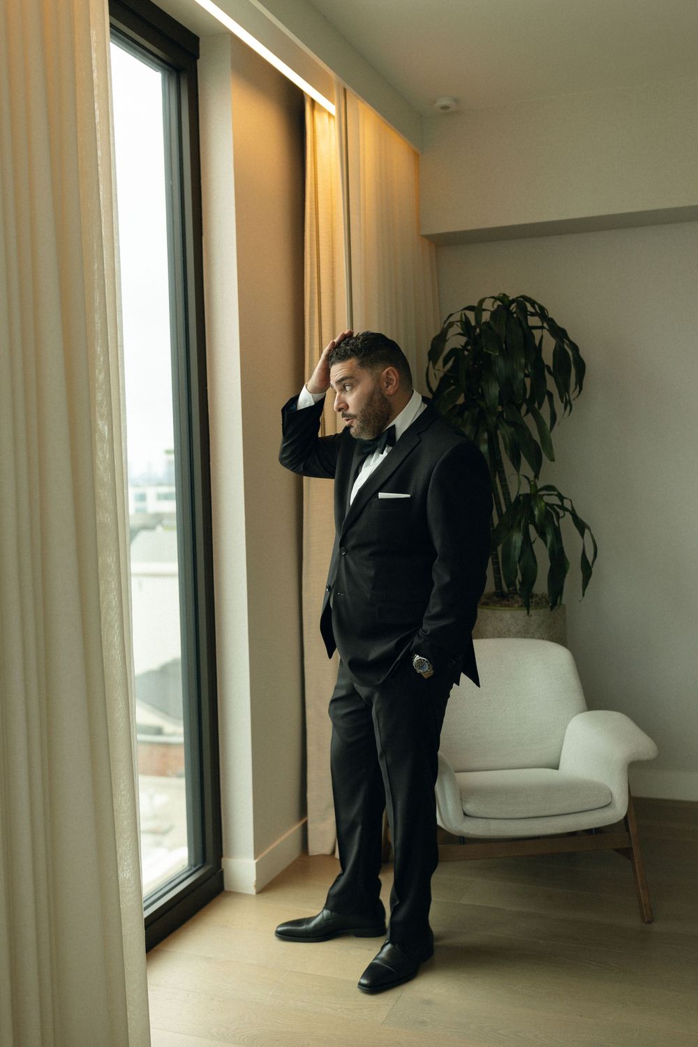 A person in a formal black tuxedo stands contemplatively by a large window in a modern room with a white armchair.