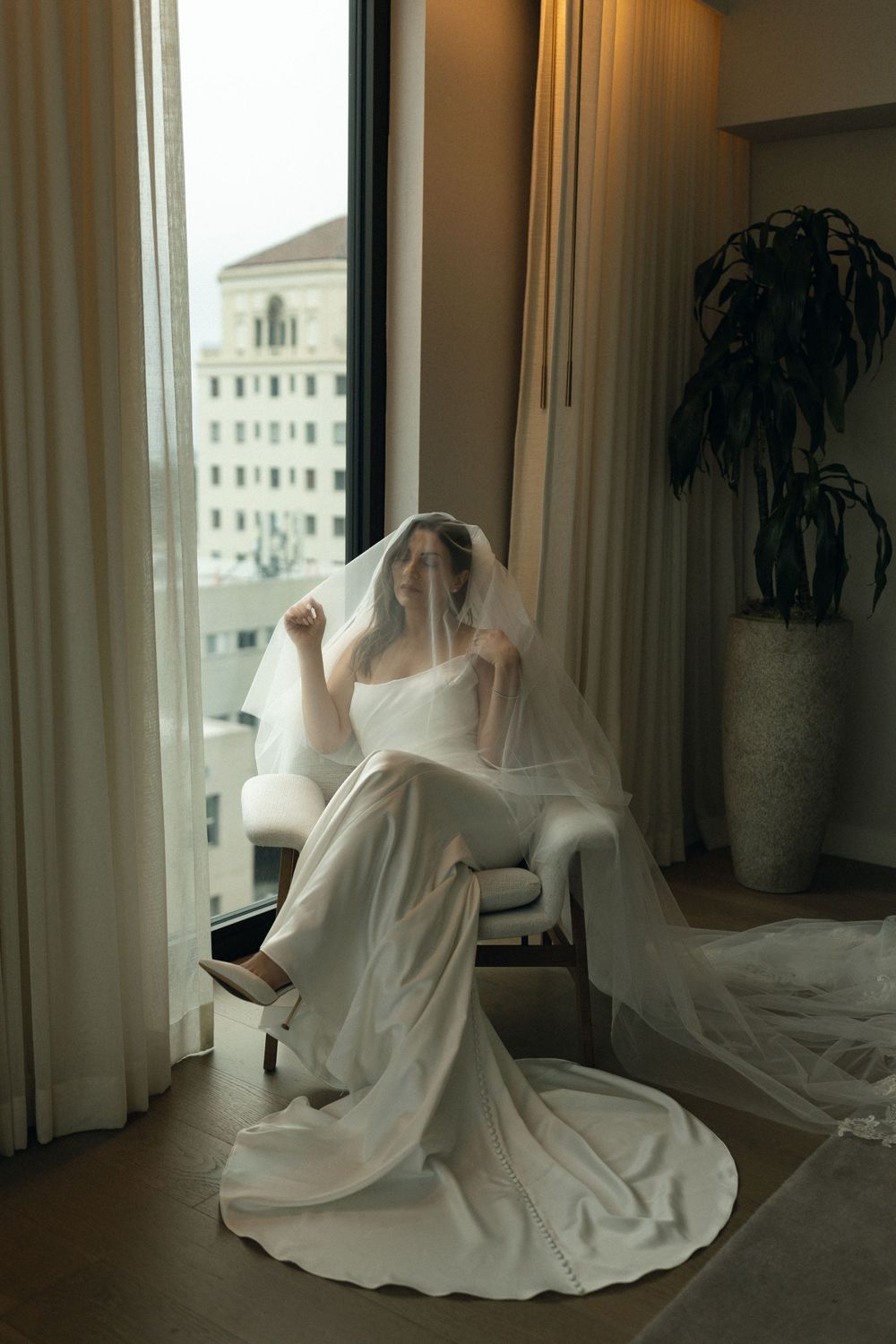 A bride in a flowing white gown and veil sits elegantly by a window with city views in a modern hotel room.