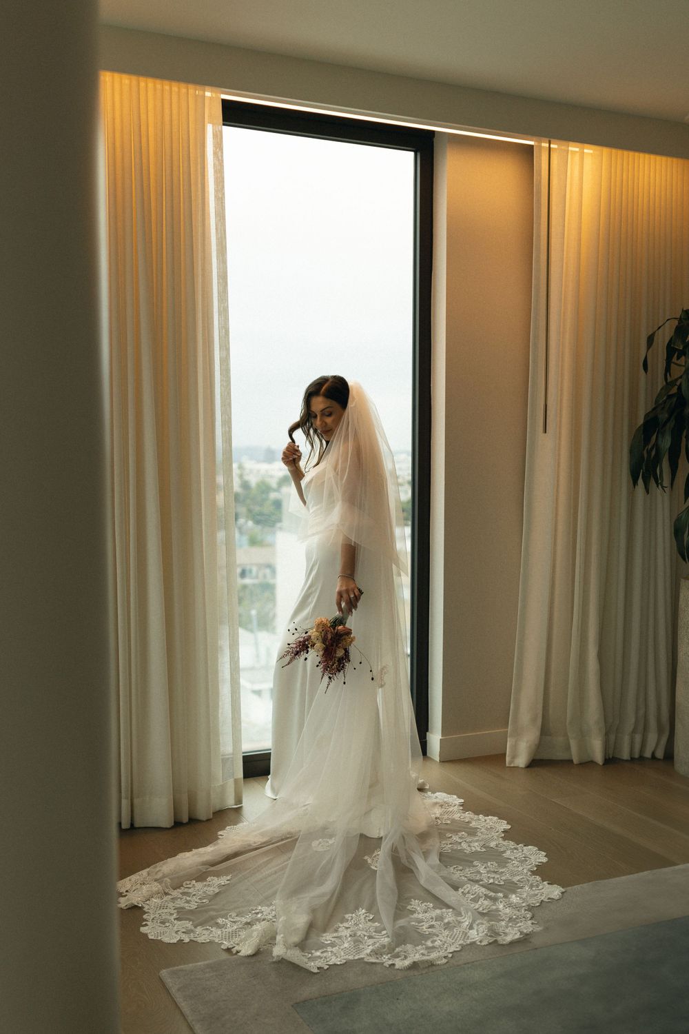 A bride in an elegant white lace wedding dress with long train and veil stands silhouetted against a bright window.