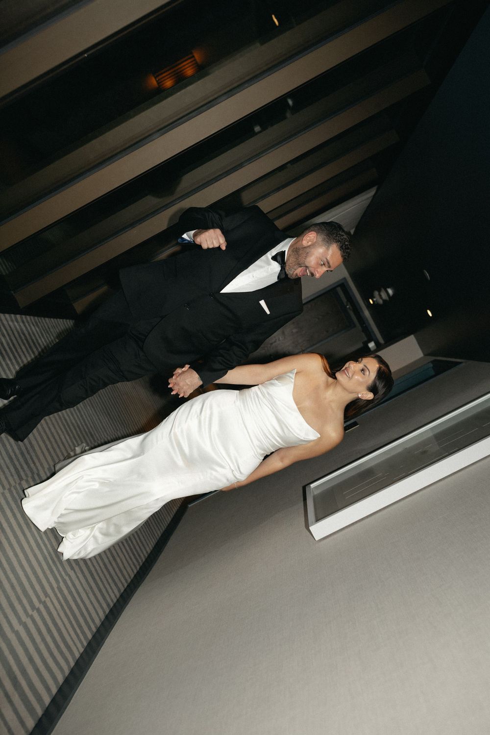 A couple in formal wedding attire pose lying down on an angular platform in a darkly lit industrial space.