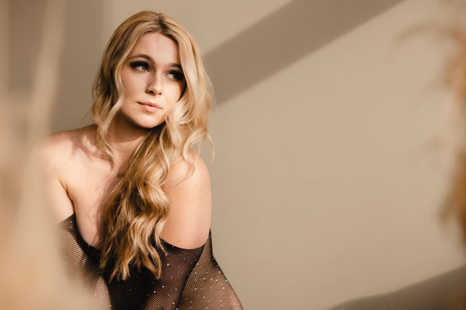 A portrait against a beige wall background features long wavy blonde hair and a black lace top in soft lighting.