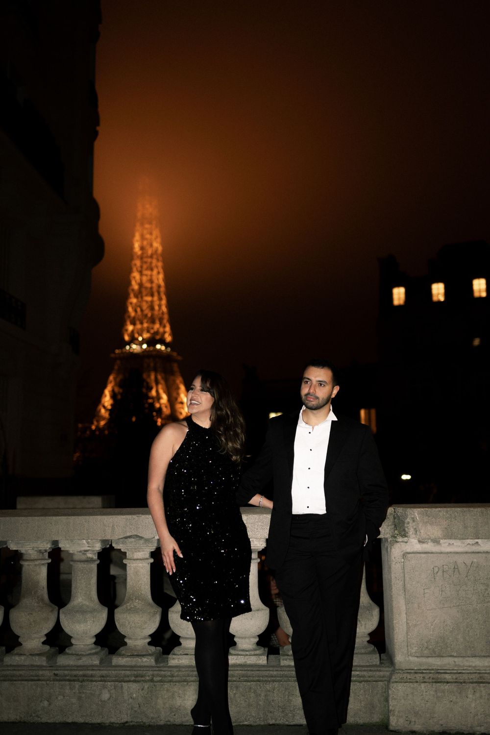 Elegant couple poses against stone balcony at night with illuminated Eiffel Tower glowing in the misty Paris backdrop.