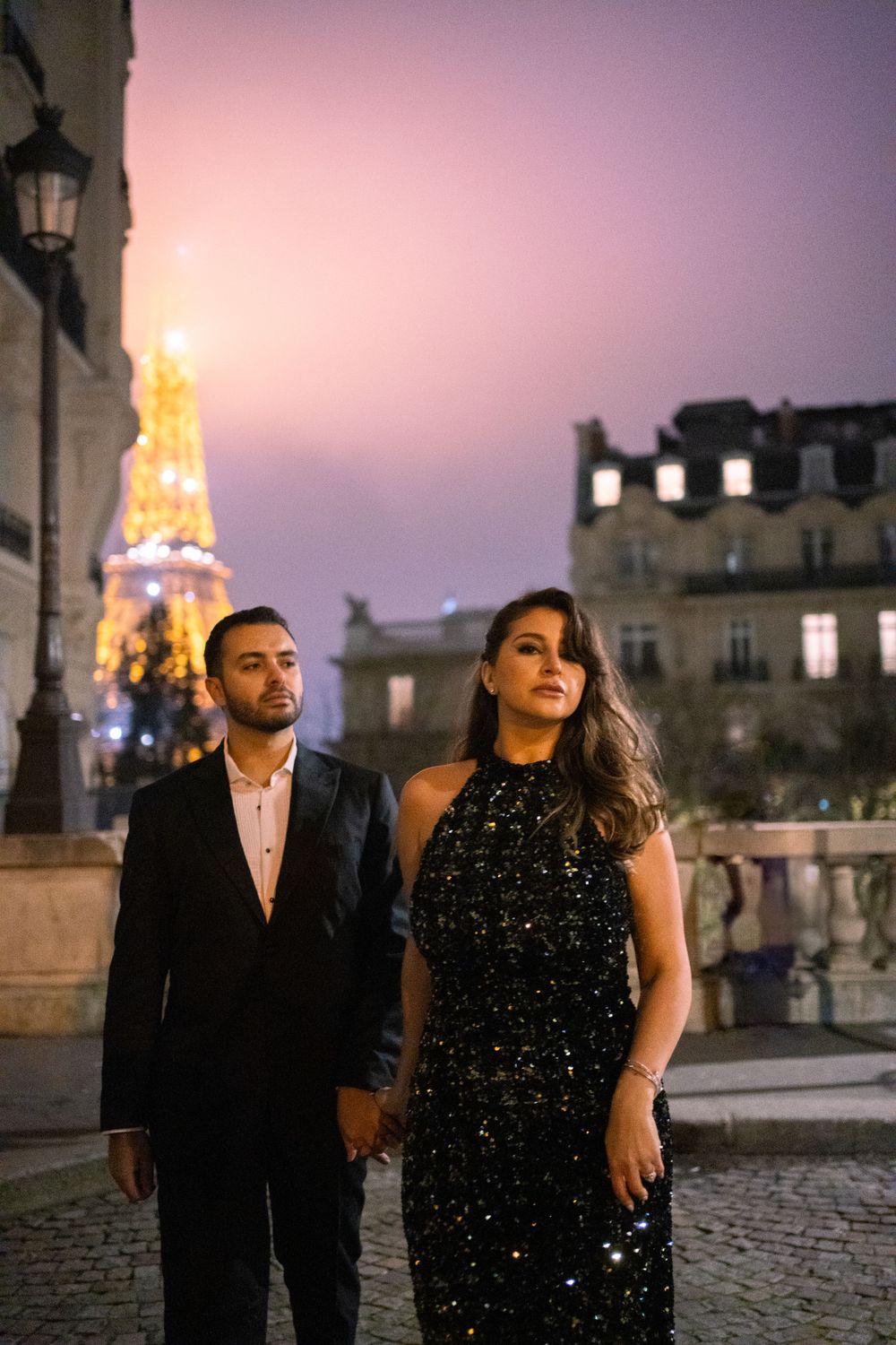 A couple in formal attire poses on a cobblestone street with the illuminated Eiffel Tower glowing in the twilight sky.
