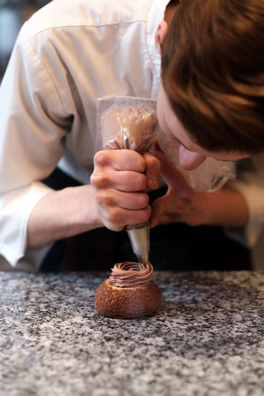 A pastry chef piping chocolate frosting from a pastry bag into a small glass jar on a granite counter.