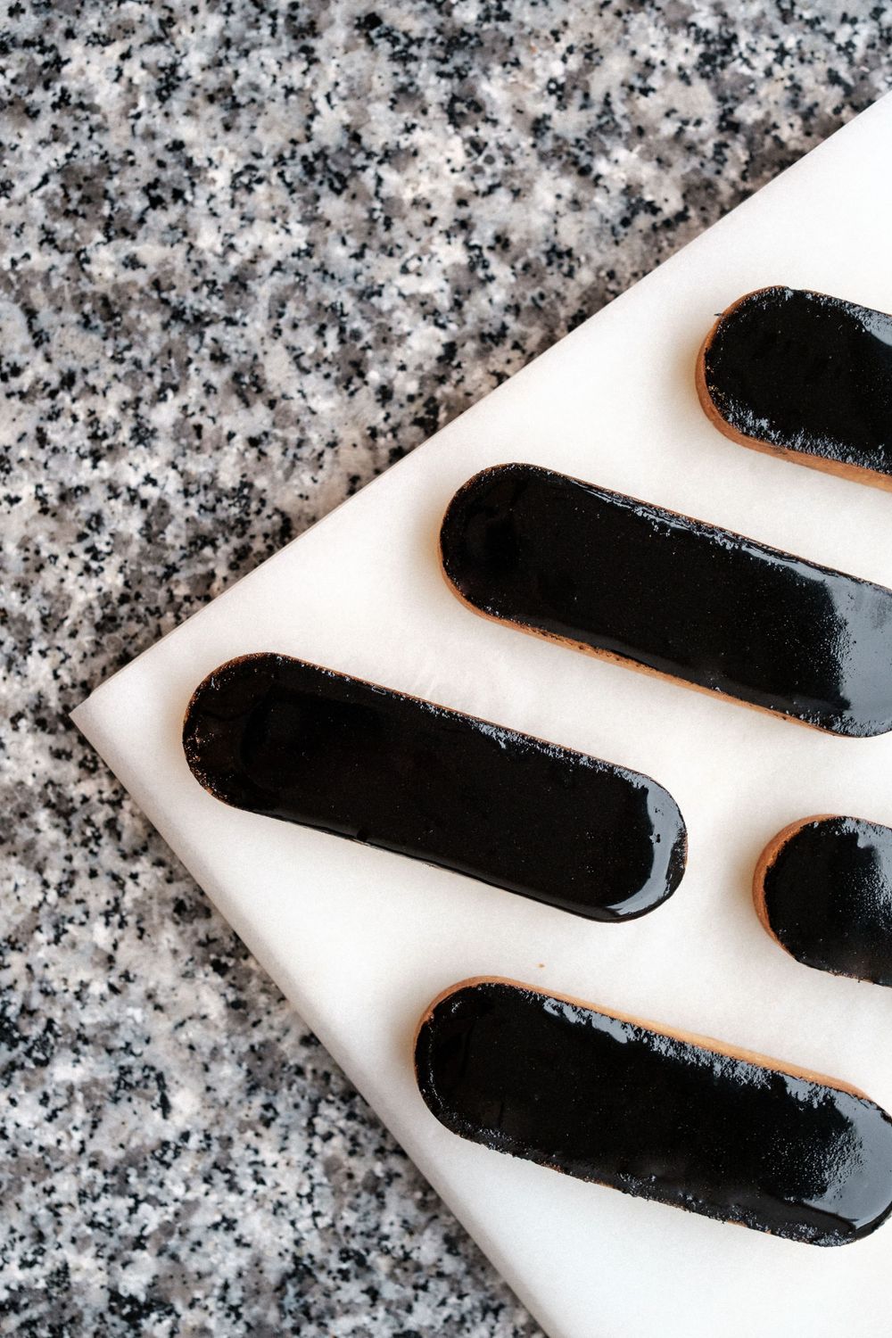 Black chocolate-covered ladyfingers on white surface next to granite countertop.