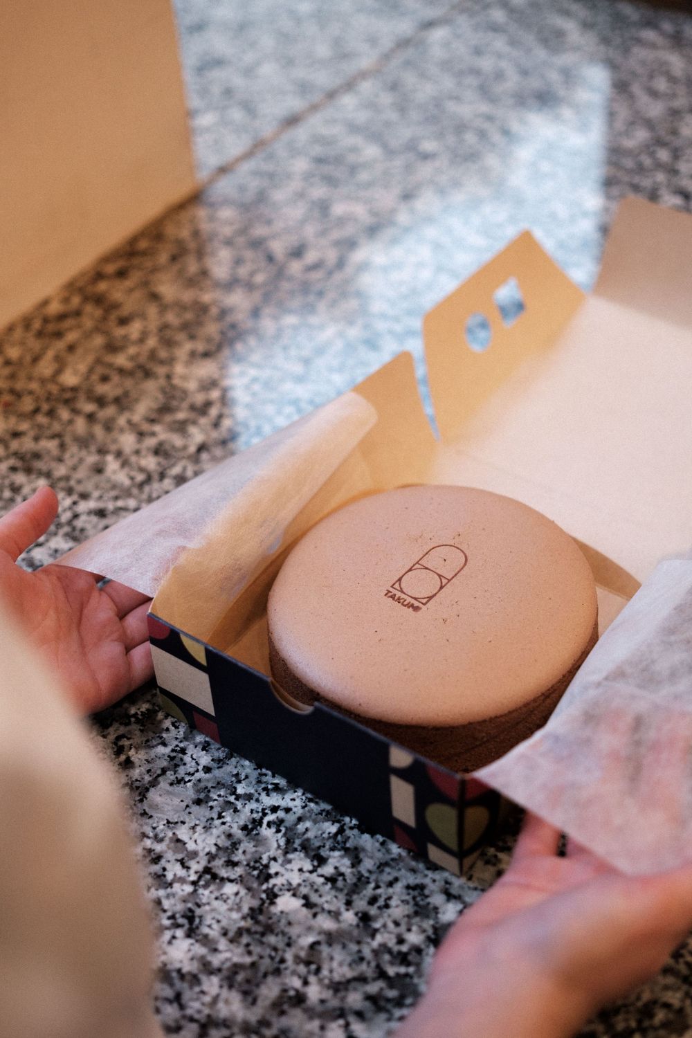 A branded pastry box being opened to reveal a circular baking mold or cake pan.
