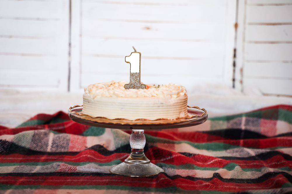 White frosted birthday cake with gold number one candle displayed on glass pedestal with plaid blanket.