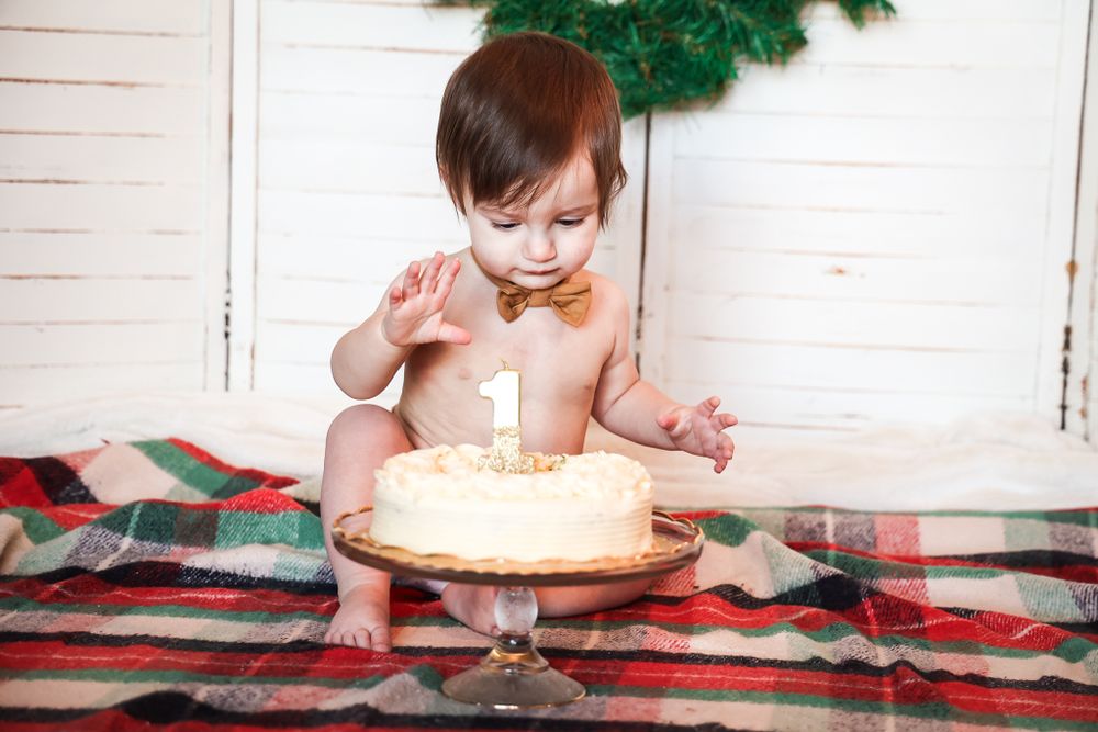 Small child reaching toward white frosted cake on glass stand with holiday decor in background.