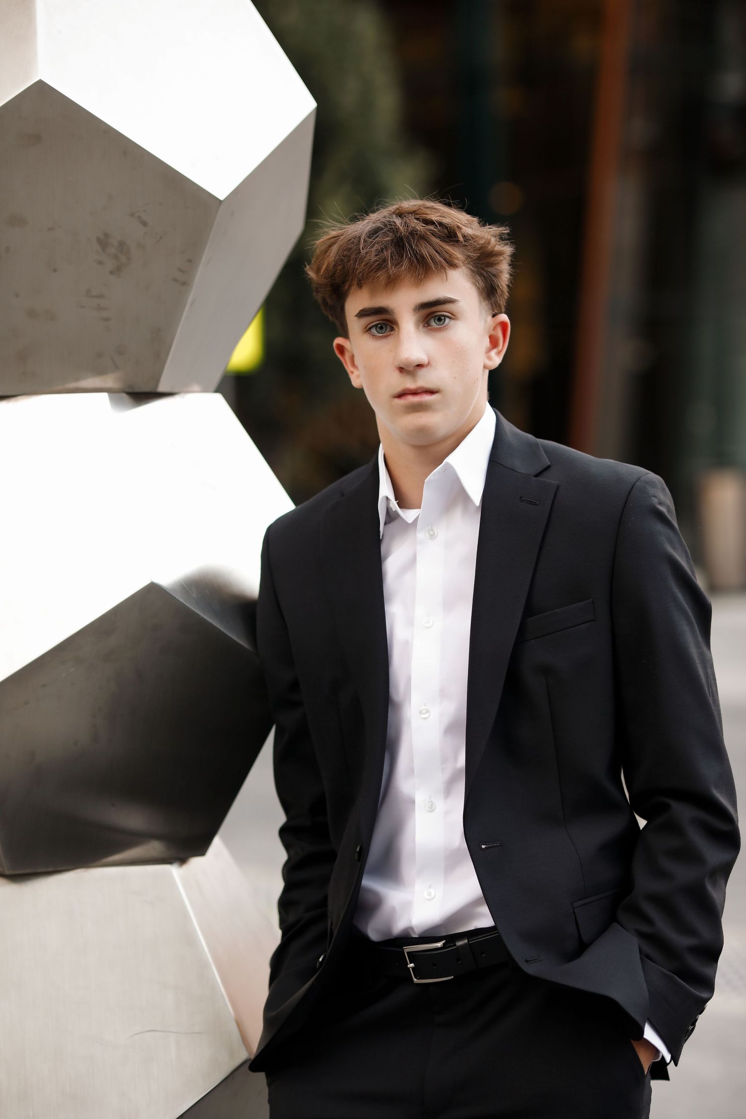 Young man in black suit and white shirt posing near geometric metal sculpture outdoors.